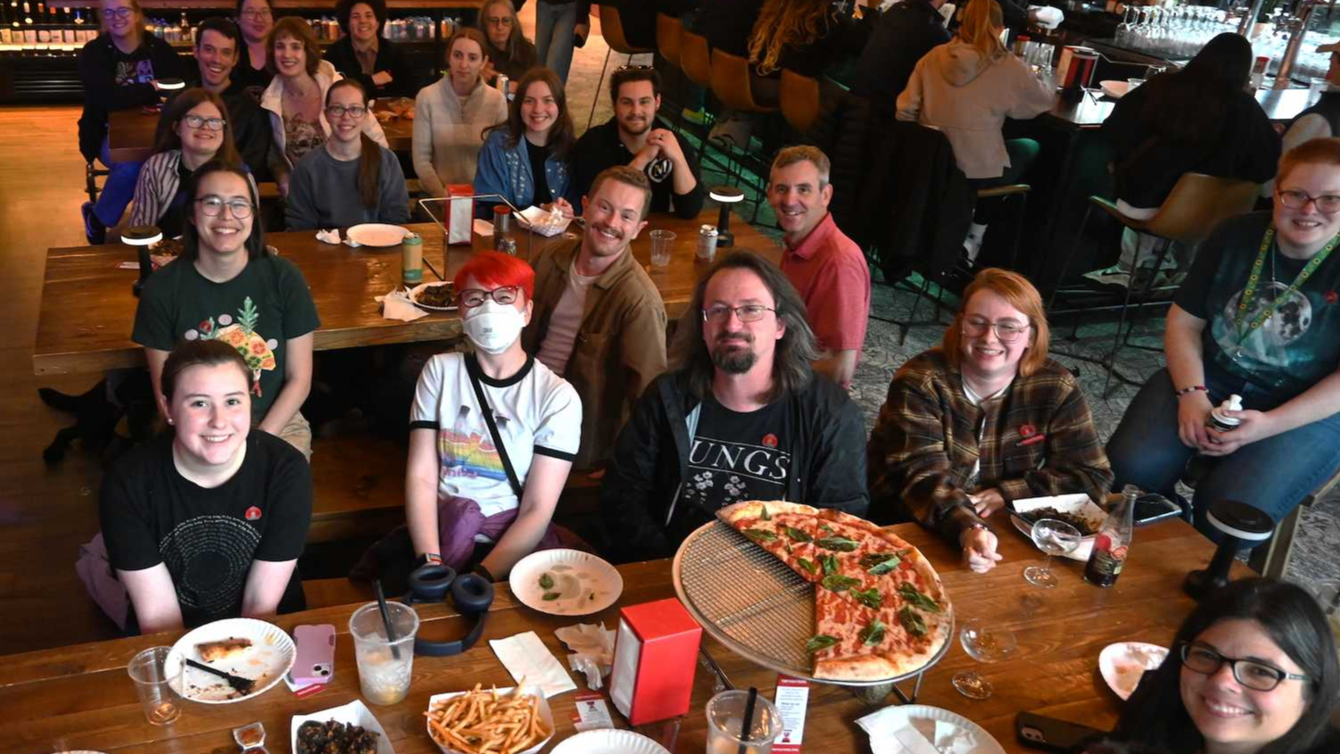 Advocates smiling at the camera wearing various nerdfighteria shirts siting at a table in a pizza place.